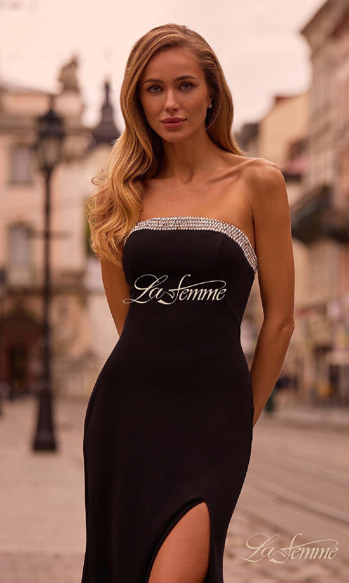 A woman wearing the Bead-Trimmed Long Prom Dress: La Femme 32891, a strapless gown with a thigh-high slit and beaded neckline, stands outdoors on a city street.